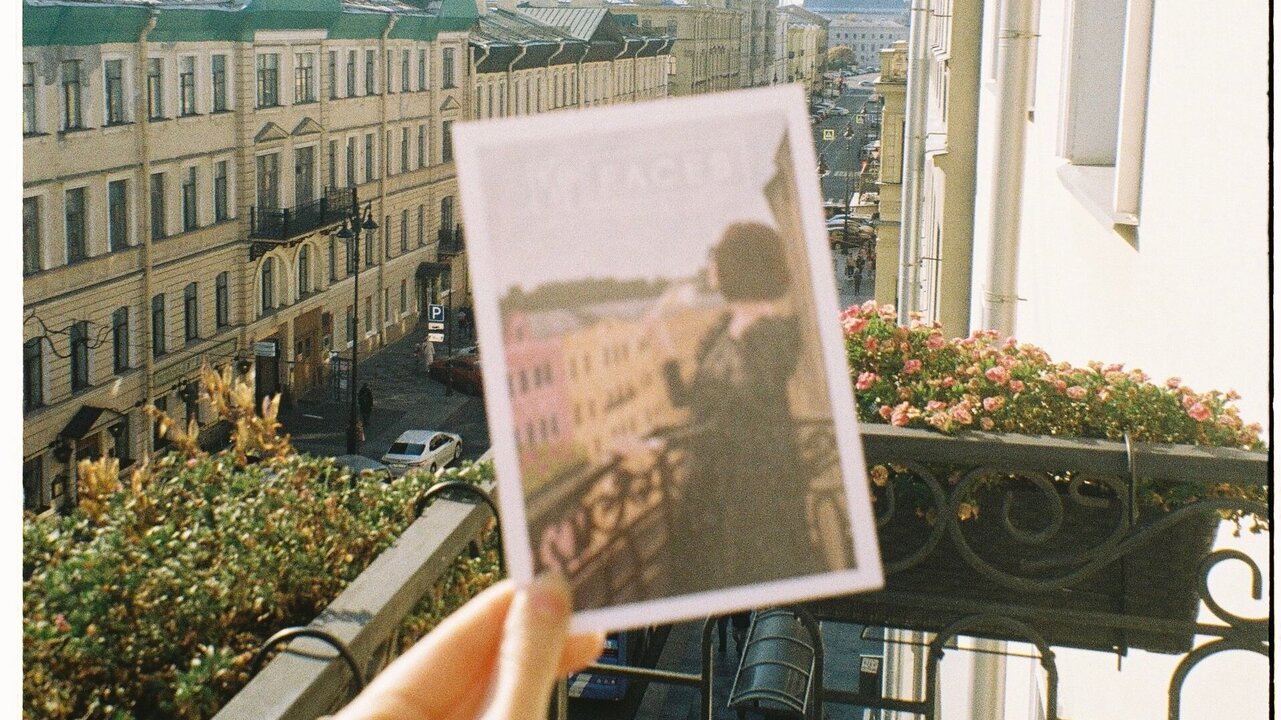 Eine Hand hält eine Postkarte mit einer Ansicht der Stadt, während sie auf einem Balkon steht. Im Hintergrund sind historische Gebäude und eine Straße sichtbar, die von einer klaren Tageslichtatmosphäre beleuchtet wird. Blumen stehen im Geländer des Balkons.