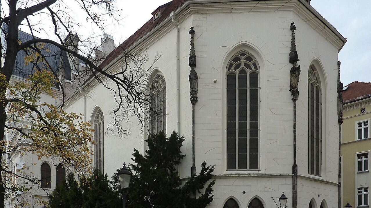 Ein historisches Kirchengebäude mit einer weißen Fassade und einem roten Satteldach. Große Fenster mit gotischen Elementen ziehen die Aufmerksamkeit auf sich. Um das Gebäude wachsen Bäume und es stehen einige Fahrräder in der Nähe.