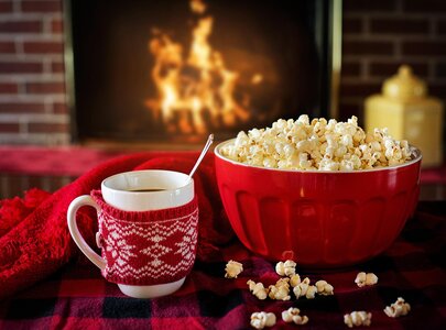 Eine Schüssel mit frisch zubereitetem Popcorn steht neben einer Tasse heißen Getränks, umwickelt mit einem roten Strickmuster. Im Hintergrund sieht man einen gemütlichen Kamin mit lodernden Flammen. Ein rotes Plaid ergänzt die warme Atmosphäre.