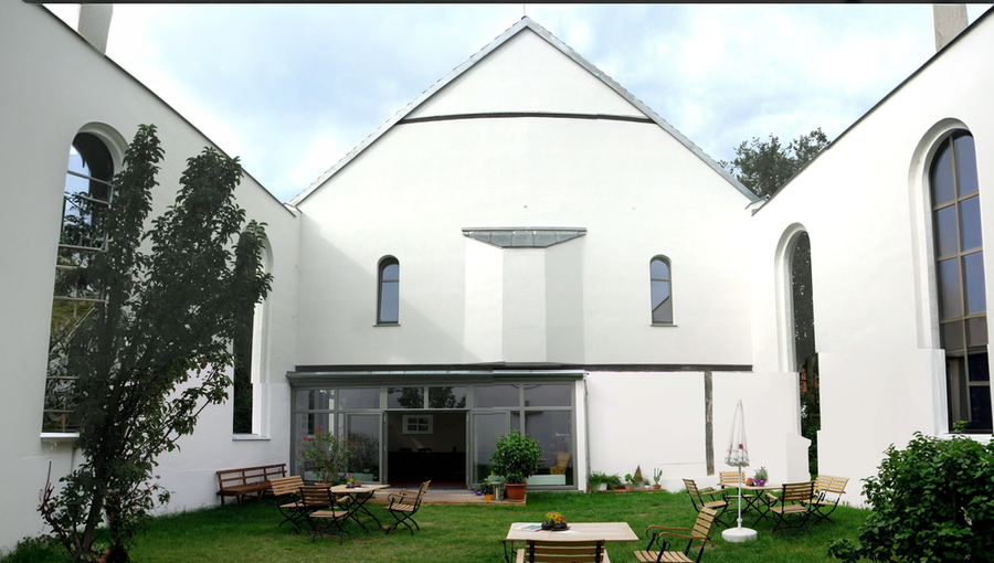 Ein heller Innenhof mit einem grünen Rasen. Umgeben von weißen Wänden und Fenstern, stehen einige Holzstühle und Tische. Im Hintergrund ist ein Glasbereich mit Pflanzen sichtbar. Die Atmosphäre wirkt einladend und ruhig.