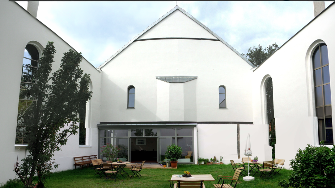 Ein heller Innenhof mit einem grünen Rasen. Umgeben von weißen Wänden und Fenstern, stehen einige Holzstühle und Tische. Im Hintergrund ist ein Glasbereich mit Pflanzen sichtbar. Die Atmosphäre wirkt einladend und ruhig.