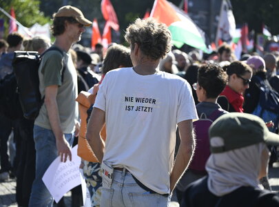 Eine Menschenmenge bei einer Demonstration. Im Vordergrund steht ein Mann mit zurückgekehrtem Kopf, der ein T-Shirt mit der Aufschrift „NIE WIEDER IST JETZT!“ trägt. Im Hintergrund sind Fahnen und weitere Teilnehmer zu sehen.