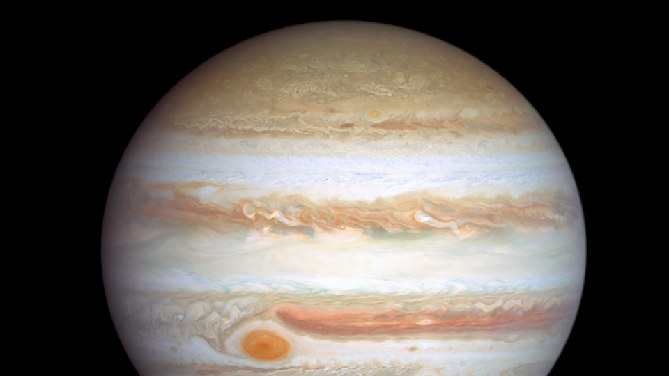 Das Bild zeigt den Planeten Jupiter mit seinen charakteristischen Streifen und dem großen roten Fleck. Neben Jupiter ist die Erde klein dargestellt. Der Hintergrund ist schwarz, was den Kontrast zwischen den Himmelskörpern verstärkt.