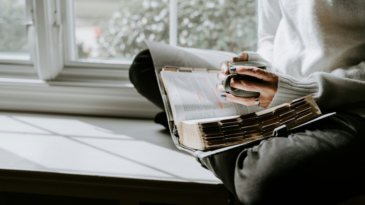 Eine Person sitzt im Schneidersitz auf einer Fensterbank und blättert durch ein Buch. Die Hände sind leicht verschmutzt von Farbe, und die Sonne scheint durch das Fenster, wodurch Schatten auf der Oberfläche entsteht.