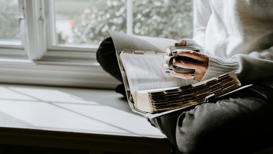 Eine Person sitzt im Schneidersitz auf einer Fensterbank und blättert durch ein Buch. Die Hände sind leicht verschmutzt von Farbe, und die Sonne scheint durch das Fenster, wodurch Schatten auf der Oberfläche entsteht.