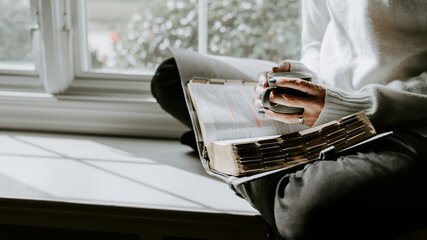 Eine Person sitzt im Schneidersitz auf einer Fensterbank und blättert durch ein Buch. Die Hände sind leicht verschmutzt von Farbe, und die Sonne scheint durch das Fenster, wodurch Schatten auf der Oberfläche entsteht.