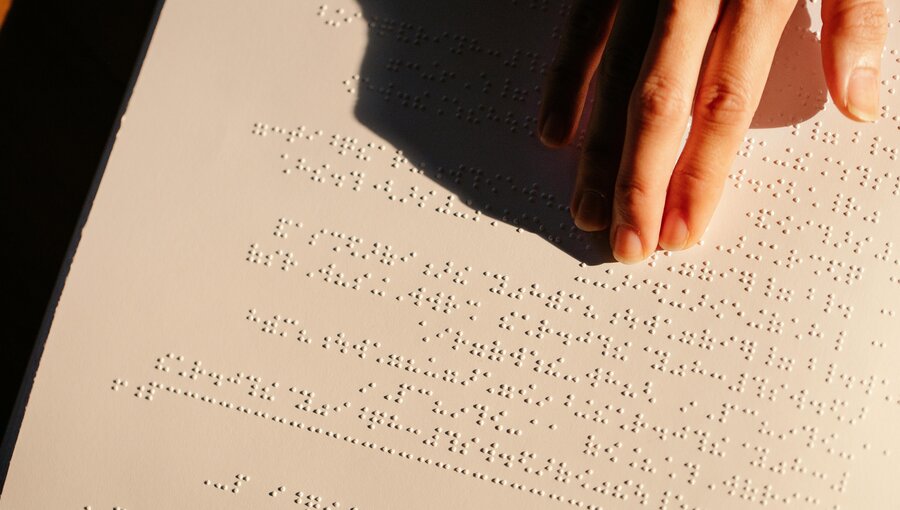Eine Hand berührt eine Oberfläche mit Blindenschrift. Die Punkte sind in Linien angeordnet, die Informationen in taktiler Form vermitteln. Die Aufnahme zeigt den Fokus auf das Lesen von Brailleschrift durch Berührung.