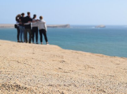 Eine Gruppe von sechs Personen steht eng zusammen an einem Sandstrand und blickt auf das Meer. Der Hintergrund zeigt das Wasser und einen klaren Himmel. Die Szene strahlt eine entspannte, freundliche Atmosphäre aus.