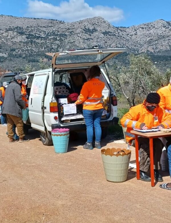 Eine Gruppe von Menschen in orangenen T-Shirts arbeitet an einem Tisch neben einem Lieferwagen. Sie stehen in einer ländlichen Umgebung mit Bergen im Hintergrund. Einige Personen sind mit Schreiben beschäftigt, während andere mit Materialien beschäftigt sind.