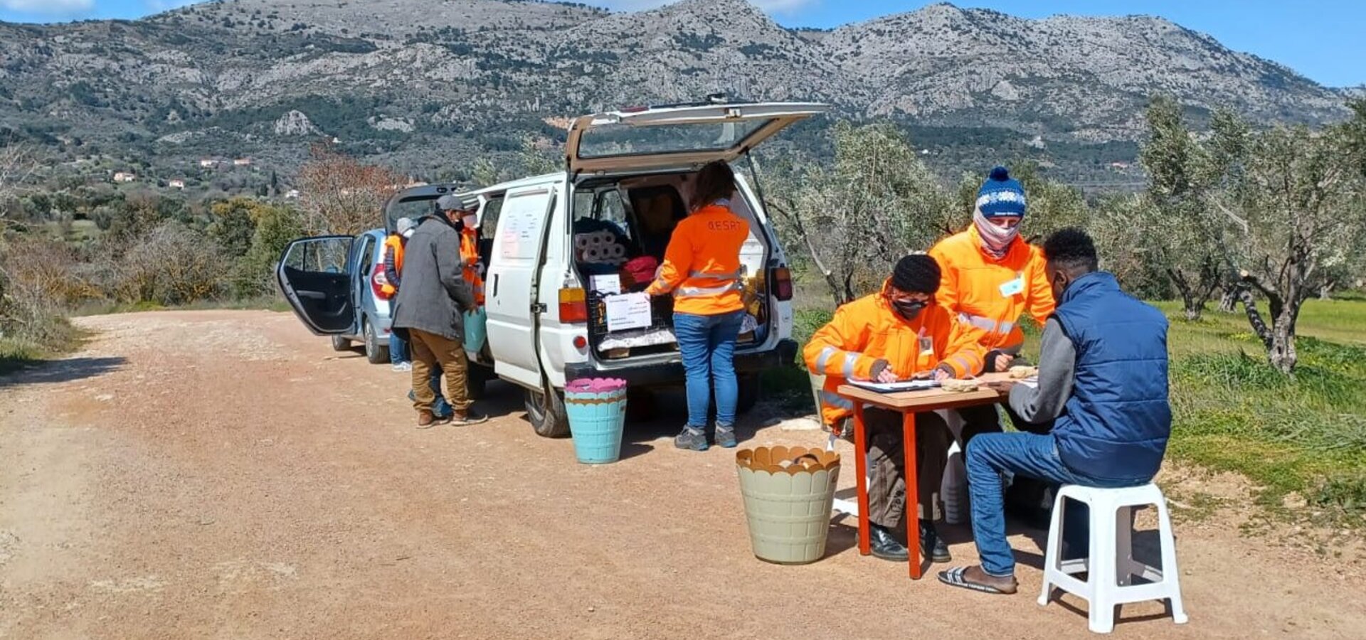Eine Gruppe von Menschen in orangenen T-Shirts arbeitet an einem Tisch neben einem Lieferwagen. Sie stehen in einer ländlichen Umgebung mit Bergen im Hintergrund. Einige Personen sind mit Schreiben beschäftigt, während andere mit Materialien beschäftigt sind.