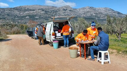 Eine Gruppe von Menschen in orangenen T-Shirts arbeitet an einem Tisch neben einem Lieferwagen. Sie stehen in einer ländlichen Umgebung mit Bergen im Hintergrund. Einige Personen sind mit Schreiben beschäftigt, während andere mit Materialien beschäftigt sind.