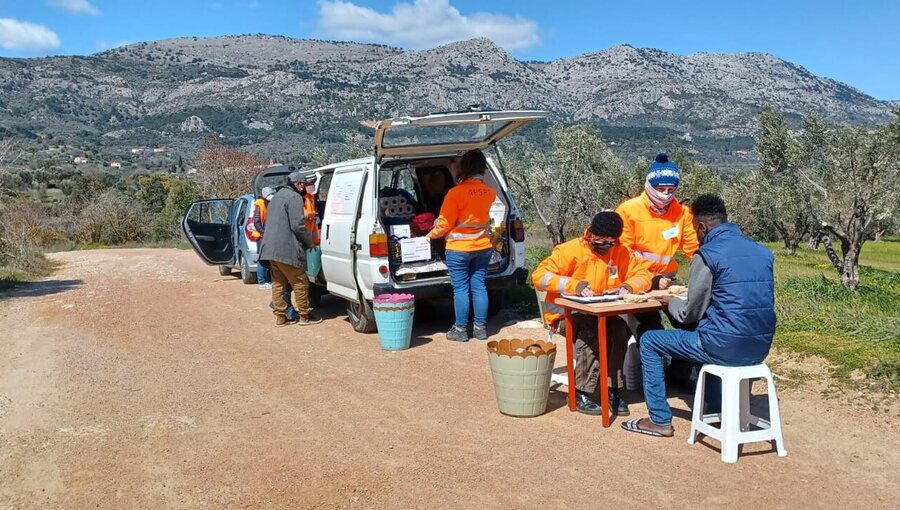 Eine Gruppe von Menschen in orangenen T-Shirts arbeitet an einem Tisch neben einem Lieferwagen. Sie stehen in einer ländlichen Umgebung mit Bergen im Hintergrund. Einige Personen sind mit Schreiben beschäftigt, während andere mit Materialien beschäftigt sind.