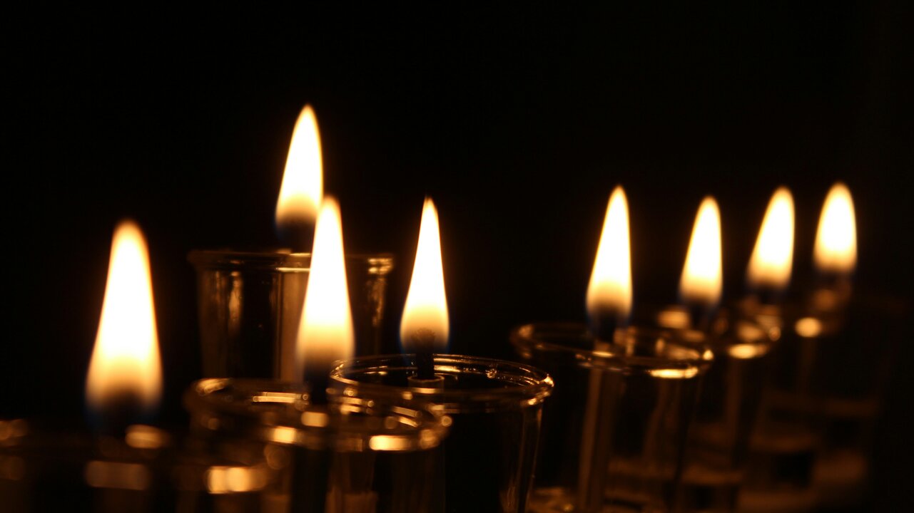 Eine Reihe von brennenden Kerzen in einem dunklen Raum. Jede Kerze hat eine flackernde Flamme, die sanft leuchtet. Der Blick ist auf die Kerzen gerichtet, die in gleichmäßigen Abständen stehen und eine warme, festliche Atmosphäre schaffen.