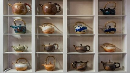 Das Bild zeigt eine Ansammlung von zwölf Teekannen, die auf einem weißen Regal angeordnet sind. Die Kannen variieren in Farbe und Design, einige sind traditionell, andere modern. Sie sind in vier Reihen zu je drei Kannen angeordnet.
