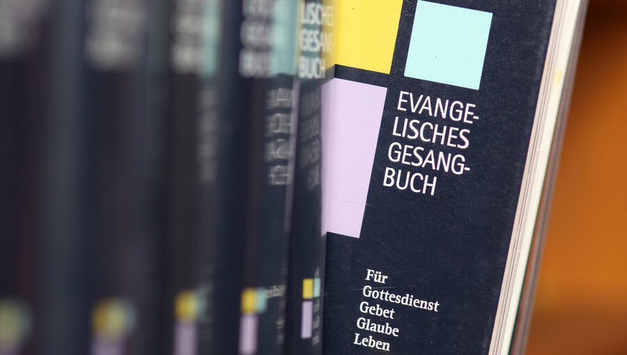 Ein evangelisches Gesangbuch hängt an einem Regal. Der Titel "Evangelisches Gesangbuch" ist deutlich sichtbar. Untertitel wie "Für Gottesdienst, Gebet, Glauben, Leben" sind zu erkennen. Farbige Bänder hängen aus dem Buch hervor, das auf eine religiöse Nutzung hinweist.