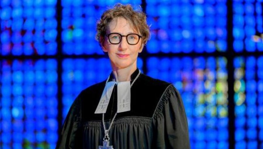 Eine lächelnde Frau trägt eine schwarze Robe und hat eine weiße Stola um den Hals. Sie steht vor einem Hintergrund aus buntem, leuchtendem Glas. Ihr Blick ist freundlich und einladend.