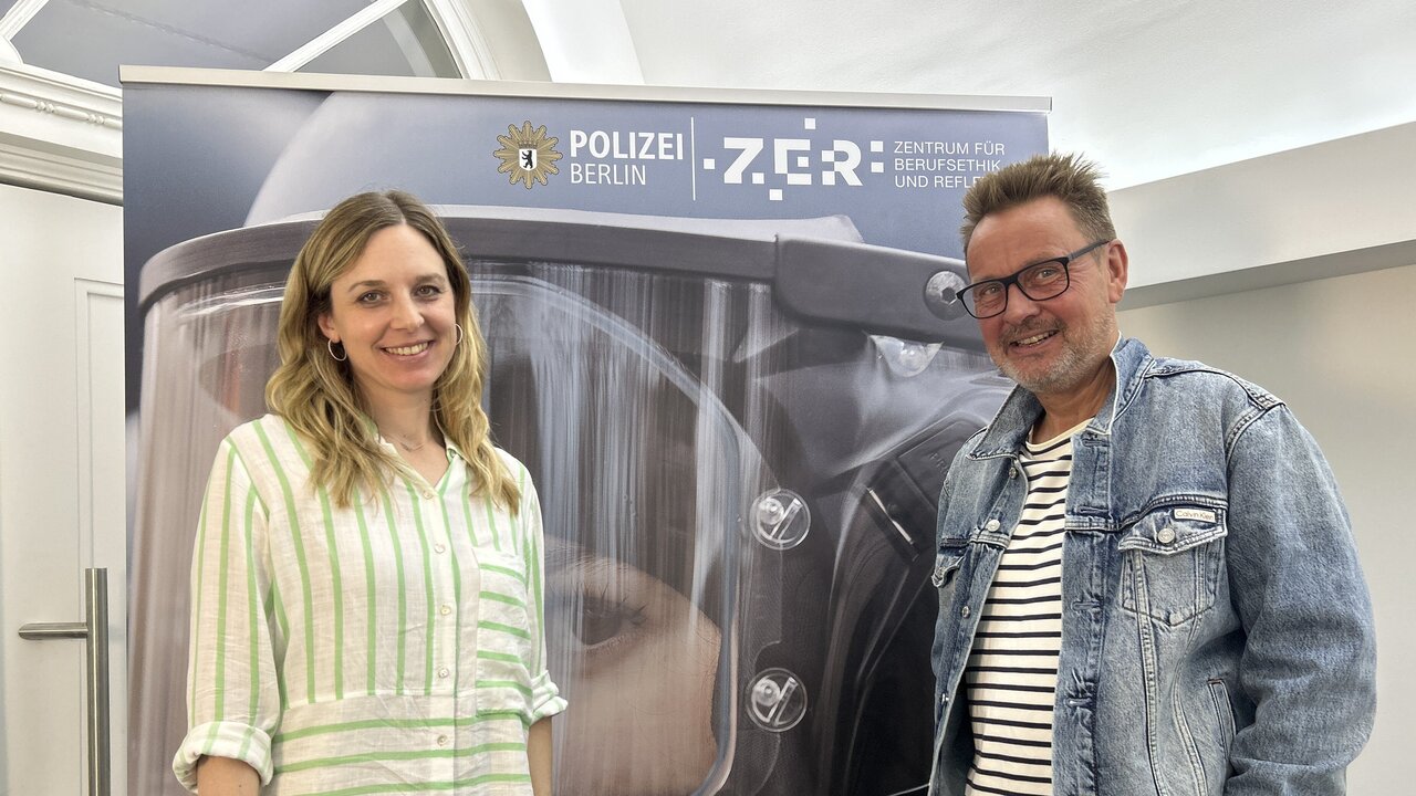 Zwei Personen stehen vor einem Hintergrund, der ein Polizeimotiv zeigt. Eine Frau trägt ein gestreiftes Hemd und lächelt, während ein Mann mit Brille und Jeansjacke neben ihr steht. Im Hintergrund ist das Logo der Polizei Berlin und der Schriftzug "ZGR - Zentrum für ...".