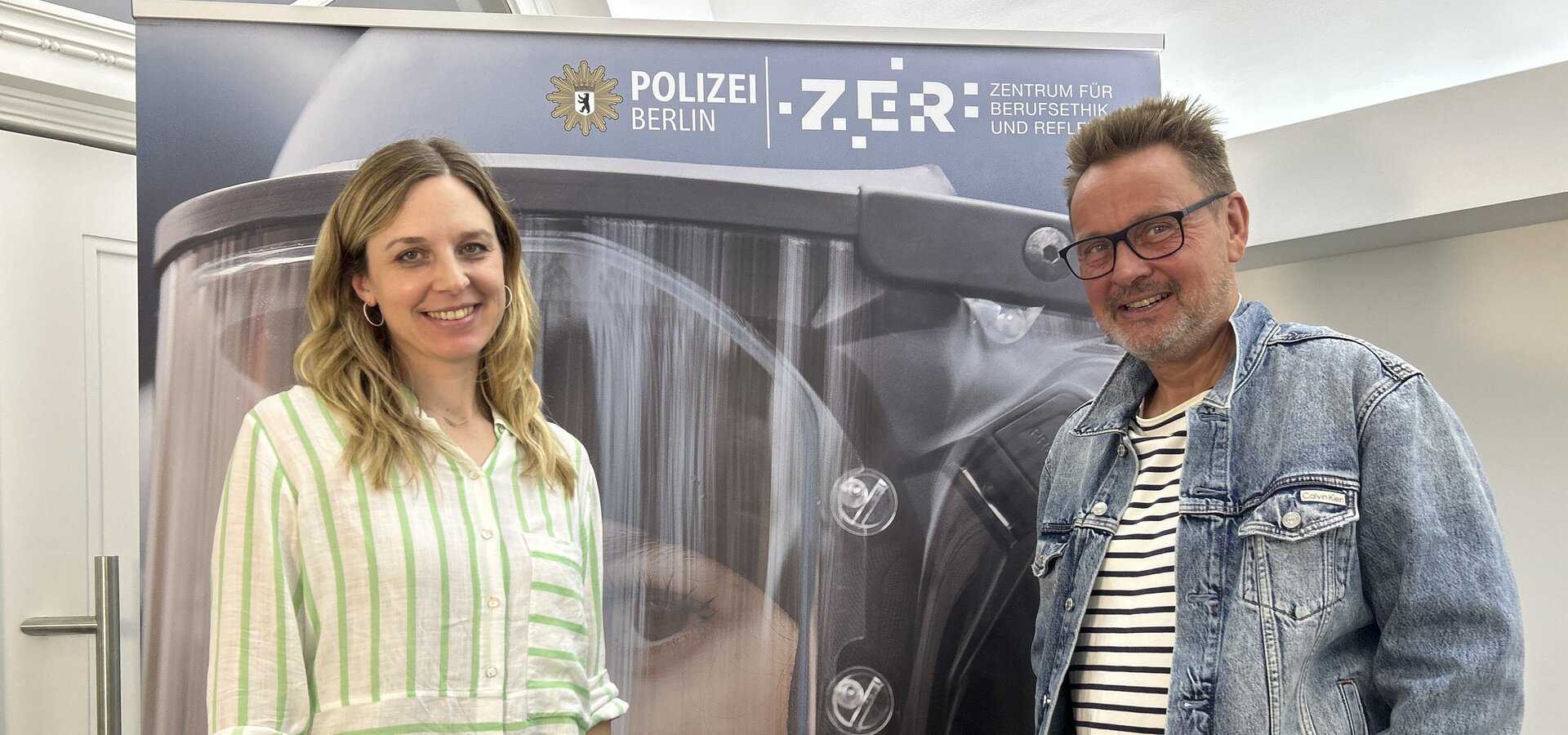 Zwei Personen stehen vor einem Hintergrund, der ein Polizeimotiv zeigt. Eine Frau trägt ein gestreiftes Hemd und lächelt, während ein Mann mit Brille und Jeansjacke neben ihr steht. Im Hintergrund ist das Logo der Polizei Berlin und der Schriftzug "ZGR - Zentrum für ...".