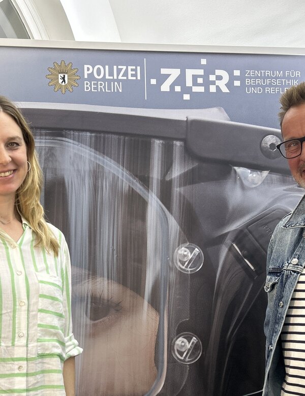 Zwei Personen stehen vor einem Hintergrund, der ein Polizeimotiv zeigt. Eine Frau trägt ein gestreiftes Hemd und lächelt, während ein Mann mit Brille und Jeansjacke neben ihr steht. Im Hintergrund ist das Logo der Polizei Berlin und der Schriftzug "ZGR - Zentrum für ...".