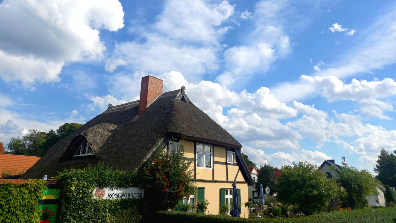Ein traditionelles Haus mit Reetdach steht unter einem blauen Himmel mit Wolken. Umgeben von Bäumen und bunten Pflanzen, vermittelt die Szene eine ruhige, ländliche Atmosphäre.
