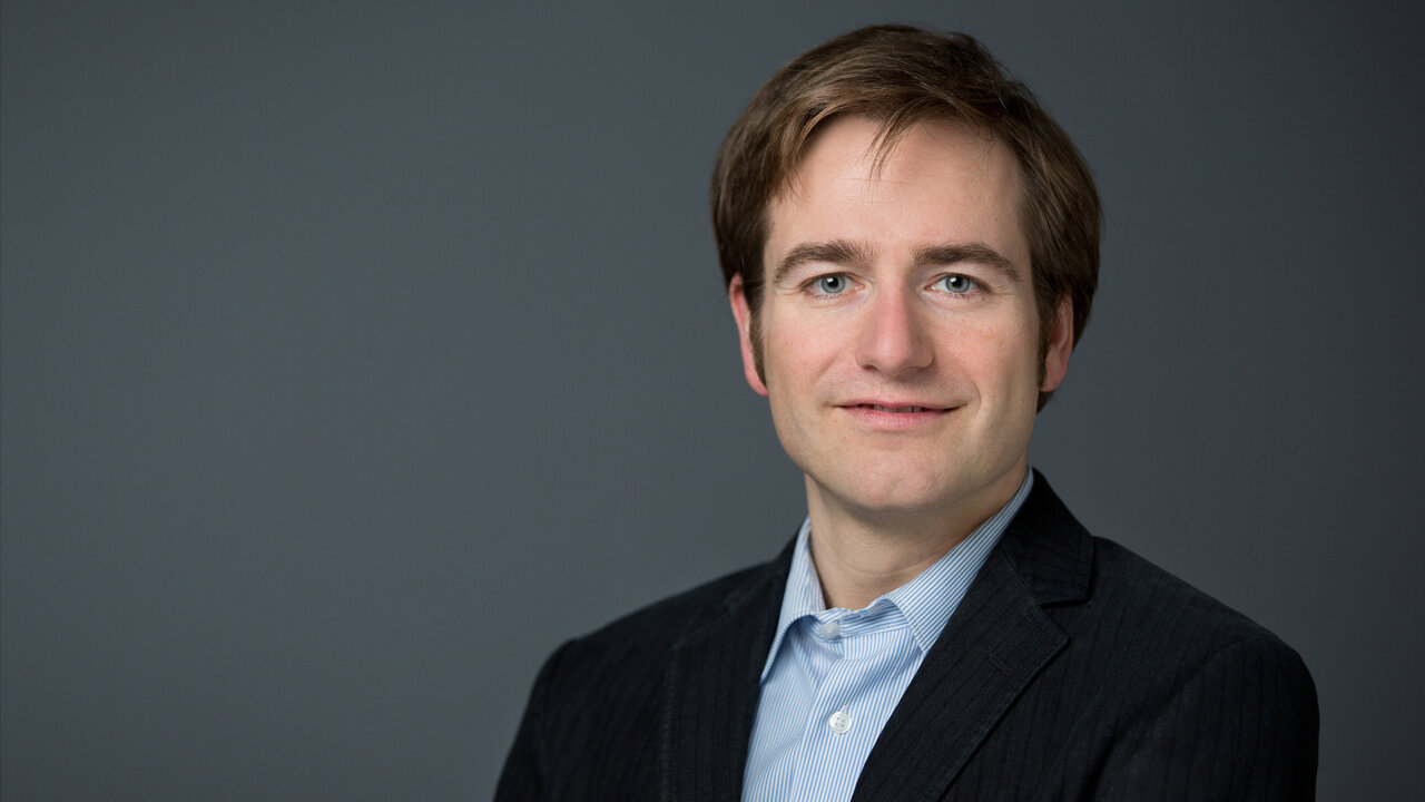 Ein lächelnder Mann mit kurzen, braunen Haaren trägt einen dunkelgrauen Anzug und ein hellblaues Hemd. Er steht vor einem neutralen, grauen Hintergrund und schaut direkt in die Kamera.