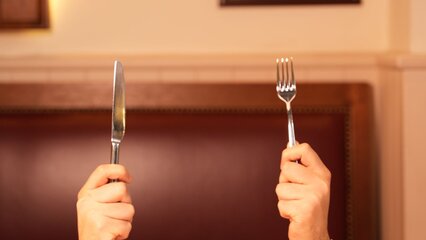 Eine Person hält mit beiden Händen ein Messer und eine Gabel, bereit, ein Stück Schokoladenkuchen mit frischen Erdbeeren und Sahne zu essen. Der Kuchen liegt auf einem Tischdecken. Im Hintergrund ist ein verschwommener, alter Mann zu sehen.