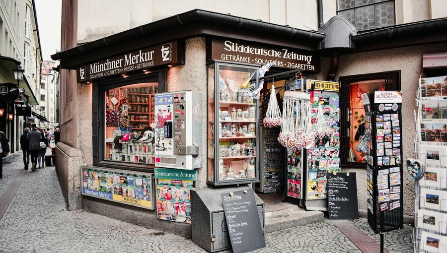 Ein Zeitungsstand in einer engen Gasse. Der Stand bietet verschiedene Zeitungen, Getränke und Souvenirs an. An der Fassade hängen Werbeschilder und Postkartenständer, während Passanten an dem Stand vorbeigehen.