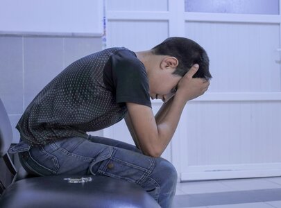 Ein junger Junge sitzt auf einem Stuhl und beugt sich mit gesenktem Kopf und den Händen im Gesicht nach vorne. Er wirkt nachdenklich und traurig, in einem Raum mit hellem Hintergrund.