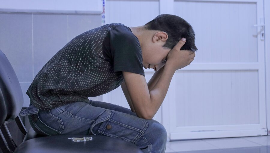 Ein junger Junge sitzt auf einem Stuhl und beugt sich mit gesenktem Kopf und den Händen im Gesicht nach vorne. Er wirkt nachdenklich und traurig, in einem Raum mit hellem Hintergrund.