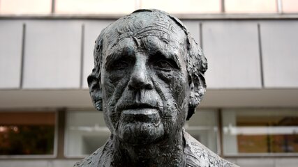 Die Skulptur zeigt ein detailliertes Porträt eines alten Mannes mit tiefen Falten und einem ernsten Ausdruck. Der Kopf ist aus Bronze gefertigt und vermittelt eine starke, nachdenkliche Präsenz. Der Hintergrund bleibt unscharf und lenkt nicht von der Figur ab.