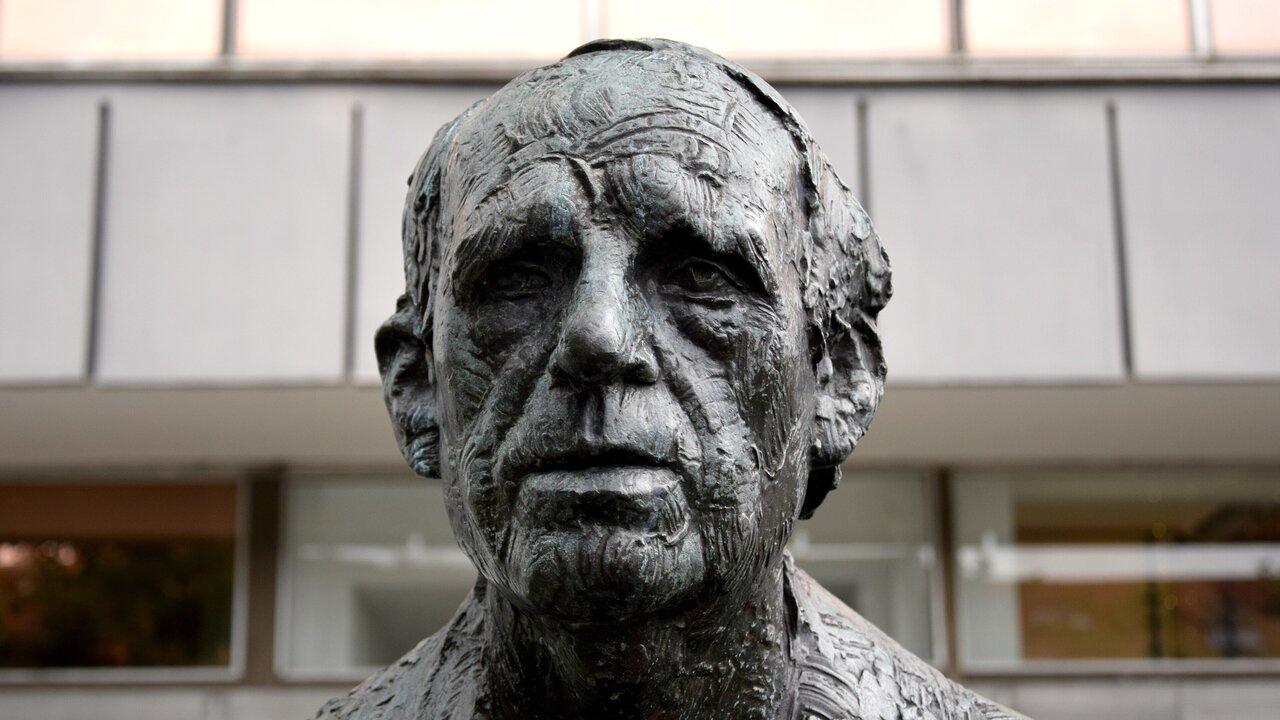 Die Skulptur zeigt ein detailliertes Porträt eines alten Mannes mit tiefen Falten und einem ernsten Ausdruck. Der Kopf ist aus Bronze gefertigt und vermittelt eine starke, nachdenkliche Präsenz. Der Hintergrund bleibt unscharf und lenkt nicht von der Figur ab.