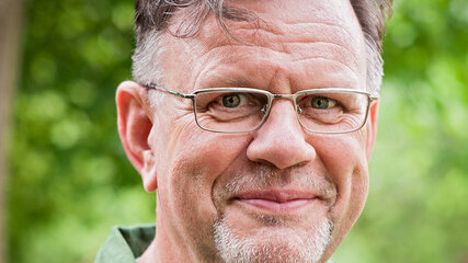 Ein lächelnder Mann mit grauen Haaren und Brille trägt ein grünes Hemd mit Mustern. Er steht im Freien vor einer grünen Kulisse aus Bäumen.