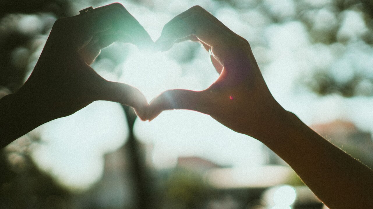 Zwei Hände formen ein Herz, während im Hintergrund helles Licht zu sehen ist. Die Silhouette der Hände hebt sich vor einem verschwommenen, natürlichen Hintergrund ab. Das Bild vermittelt ein Gefühl von Liebe und Zuneigung.