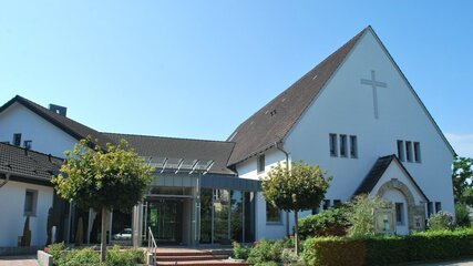 Ein modernes Kirchengebäude mit einem hohen Satteldach und einem Kreuz an der Fassade. Vor dem Hauptportal befinden sich Stufen und eine Glasüberdachung. Es gibt grüne Bäume und Sträucher, die das Gebäude umgeben, was eine einladende Atmosphäre schafft.