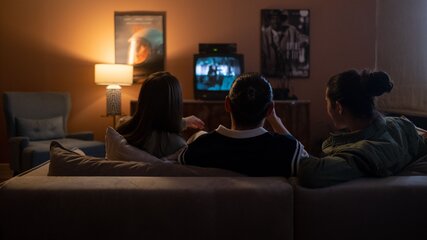 Drei Personen sitzen auf einem Sofa und schauen gemeinsam einen Fernseher an. Der Raum ist schwach beleuchtet, eine Lampe spendet Licht. Im Hintergrund sind Poster an der Wand zu sehen. Die Stimmung ist entspannt und familiär.