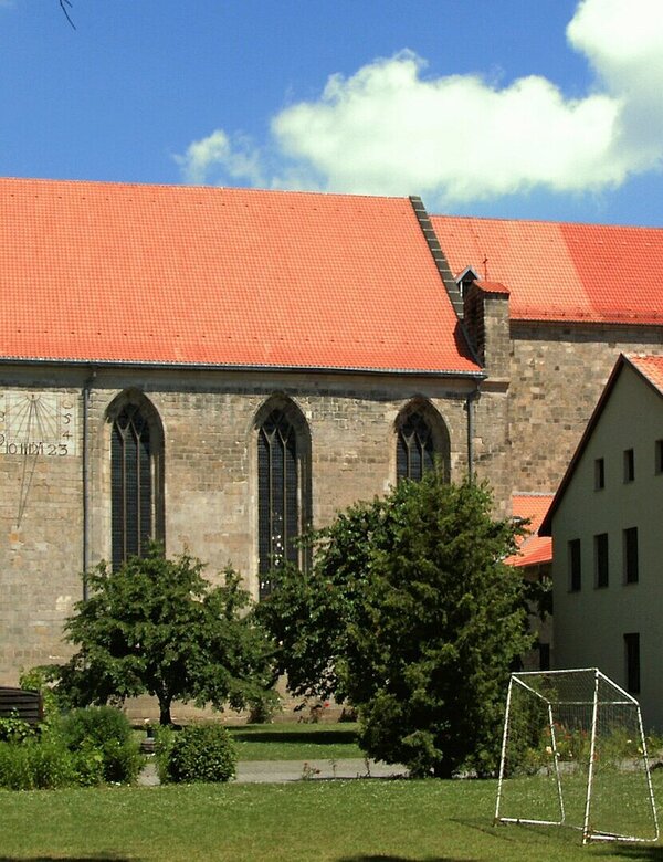 Eine Kirche mit rotem Dach steht im Vordergrund, flankiert von Bäumen. Daneben befindet sich ein Gebäude mit ebenfalls rotem Dach. Im Vordergrund ist eine Wiese mit einem Fußballtor zu sehen. Der Himmel ist blau mit einigen weißen Wolken.