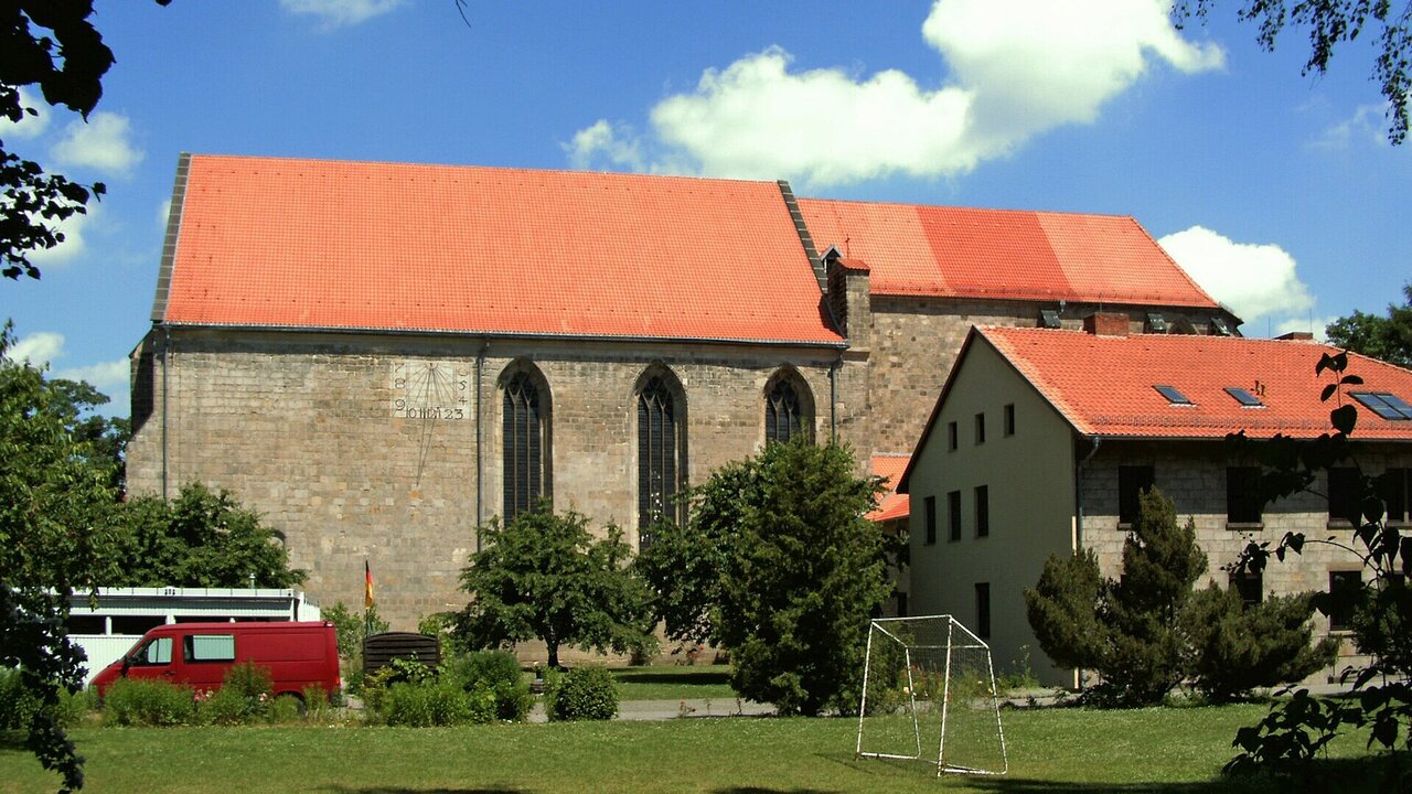 Eine Kirche mit rotem Dach steht im Vordergrund, flankiert von Bäumen. Daneben befindet sich ein Gebäude mit ebenfalls rotem Dach. Im Vordergrund ist eine Wiese mit einem Fußballtor zu sehen. Der Himmel ist blau mit einigen weißen Wolken.