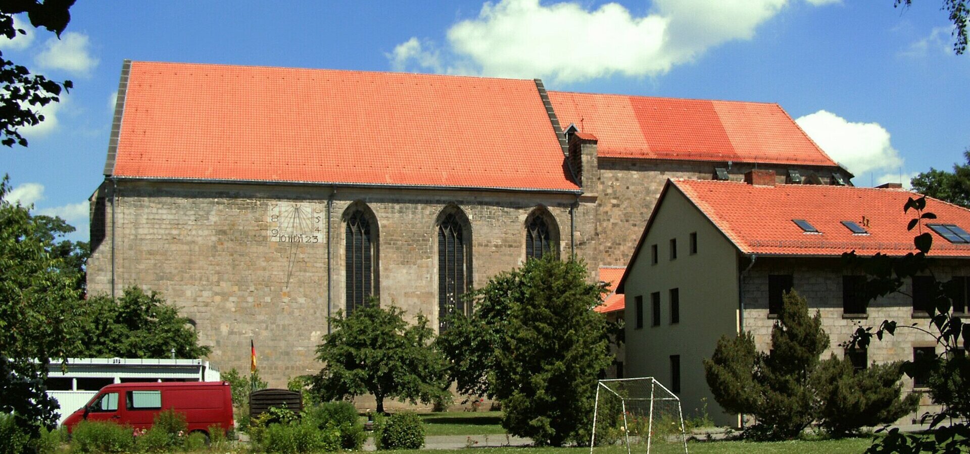 Eine Kirche mit rotem Dach steht im Vordergrund, flankiert von Bäumen. Daneben befindet sich ein Gebäude mit ebenfalls rotem Dach. Im Vordergrund ist eine Wiese mit einem Fußballtor zu sehen. Der Himmel ist blau mit einigen weißen Wolken.