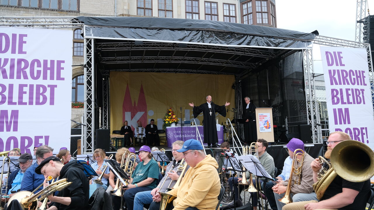 Eine Bühne mit mehreren Musikern, die Blasinstrumente spielen. Im Hintergrund steht ein Redner mit ausgebreiteten Armen, während eine andere Person neben ihm spricht. Große Banner verkünden: "DIE KIRCHE BLEIBT IM MOORE!" und es sind Menschen versammelt.