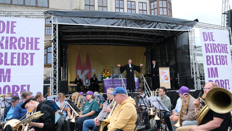 Eine Bühne mit mehreren Musikern, die Blasinstrumente spielen. Im Hintergrund steht ein Redner mit ausgebreiteten Armen, während eine andere Person neben ihm spricht. Große Banner verkünden: "DIE KIRCHE BLEIBT IM MOORE!" und es sind Menschen versammelt.