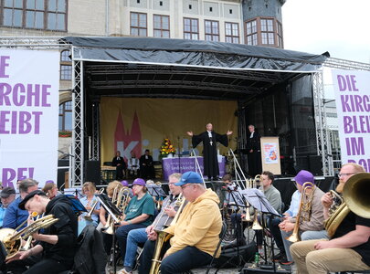 Eine Bühne mit mehreren Musikern, die Blasinstrumente spielen. Im Hintergrund steht ein Redner mit ausgebreiteten Armen, während eine andere Person neben ihm spricht. Große Banner verkünden: "DIE KIRCHE BLEIBT IM MOORE!" und es sind Menschen versammelt.