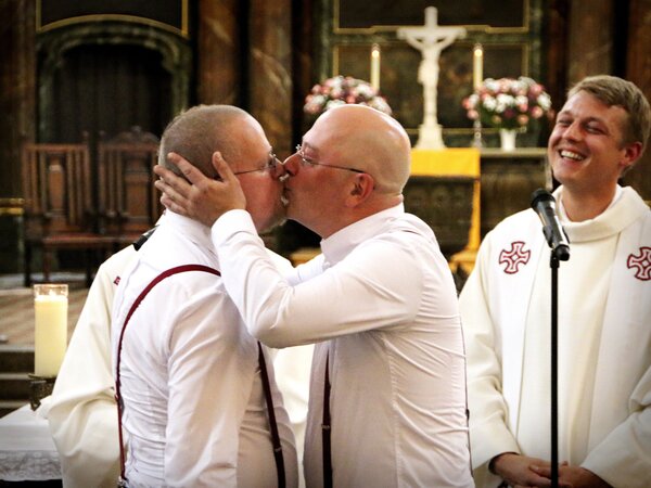 Ein Brautpaar in formeller Kleidung küsst sich in einer Kirche während der Zeremonie. Im Hintergrund steht ein Geistlicher lächelnd mit einem Mikrofon. Die Umgebung ist festlich dekoriert, was eine feierliche Stimmung vermittelt.