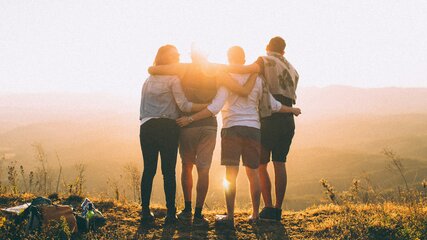 Vier Personen stehen Rücken an Rücken auf einem Hügel und betrachten den Sonnenuntergang. Sie umarmen sich und schauen in die Ferne, während warmes Licht die Szene erleuchtet. Die Landschaft hinter ihnen ist hügelig und bewaldet.