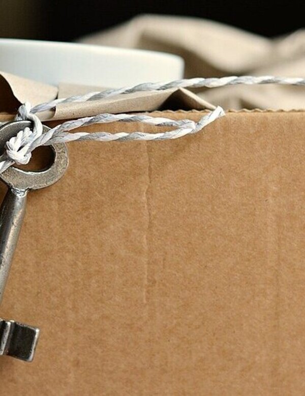 Ein Schlüssel hängt an einer Schnur, die an einer braunen Kartonbox befestigt ist. Die Box scheint mit einer Tasse und weiteren Gegenständen gefüllt zu sein. Der Schlüssel deutet darauf hin, dass die Box möglicherweise eine besondere oder geschützte Ware enthält.