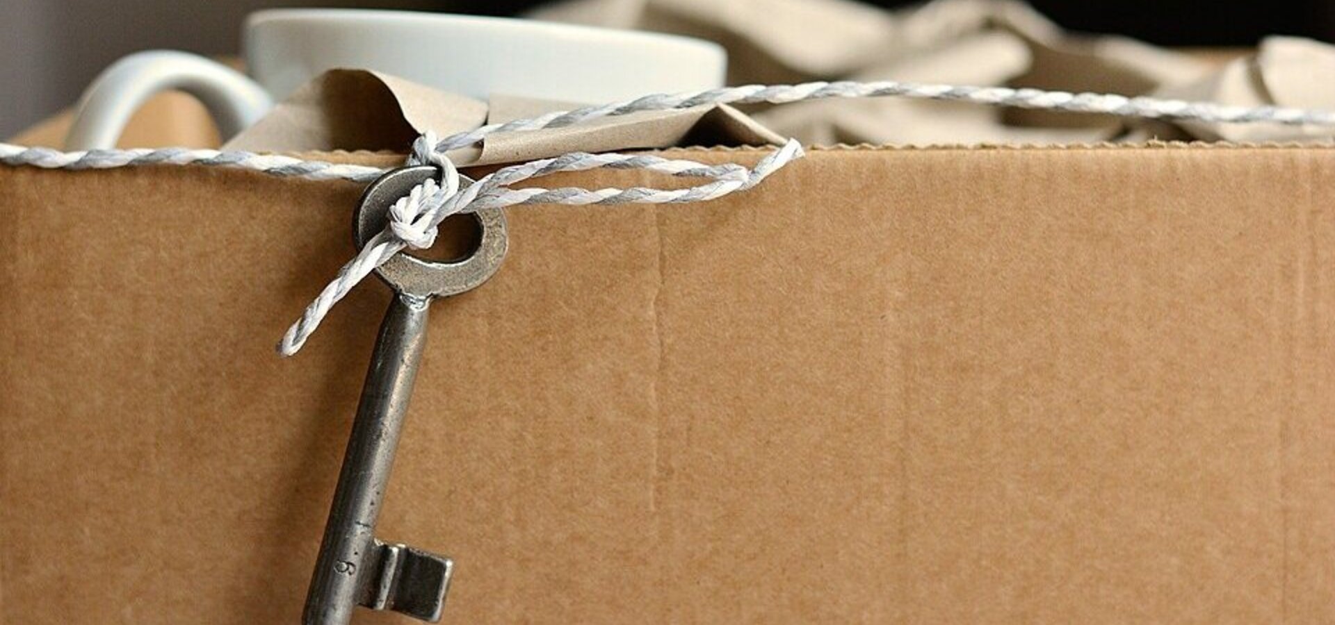 Ein Schlüssel hängt an einer Schnur, die an einer braunen Kartonbox befestigt ist. Die Box scheint mit einer Tasse und weiteren Gegenständen gefüllt zu sein. Der Schlüssel deutet darauf hin, dass die Box möglicherweise eine besondere oder geschützte Ware enthält.