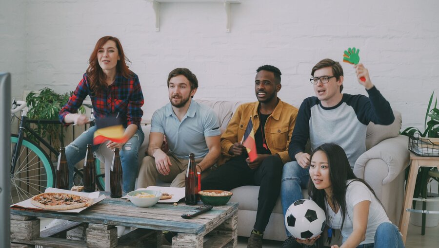 Eine Gruppe von fünf Menschen sitzt auf einem Sofa und schaut fassungsvoll einen Fernseher, während sie ein Fußballspiel verfolgen. Sie haben kleine Flaggen in der Hand und es gibt Snacks auf dem Tisch. Die Stimmung ist begeistert und familiär.