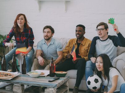 Eine Gruppe von fünf Menschen sitzt auf einem Sofa und schaut fassungsvoll einen Fernseher, während sie ein Fußballspiel verfolgen. Sie haben kleine Flaggen in der Hand und es gibt Snacks auf dem Tisch. Die Stimmung ist begeistert und familiär.