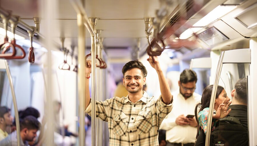 Ein junger Mann mit einem freundlichen Lächeln steht in einer U-Bahn, hält sich an einem Haltegriff fest. Im Hintergrund sind weitere Fahrgäste zu sehen, die in ihre Smartphones schauen oder miteinander sprechen. Die Atmosphäre ist lebhaft und urban.