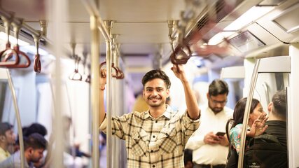 Ein junger Mann mit einem freundlichen Lächeln steht in einer U-Bahn, hält sich an einem Haltegriff fest. Im Hintergrund sind weitere Fahrgäste zu sehen, die in ihre Smartphones schauen oder miteinander sprechen. Die Atmosphäre ist lebhaft und urban.