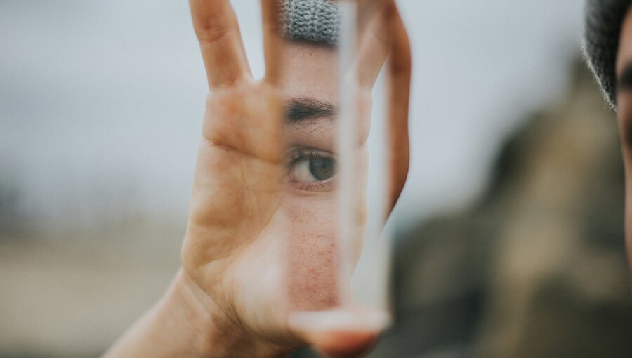 Eine Hand hält ein prismatisches Glas vor ein Auge. Durch das Glas ist das Auge deutlich sichtbar, während der Hintergrund unscharf bleibt. Die Atmosphäre vermittelt einen Eindruck von Reflektion und introspektiver Betrachtung.